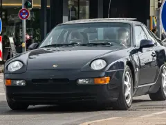 Dunkelblauer 968 Porsche fährt auf der Straße. Ein Mann sitzt am Steuer. Verkehrsschilder sind im Hintergrund zu sehen.