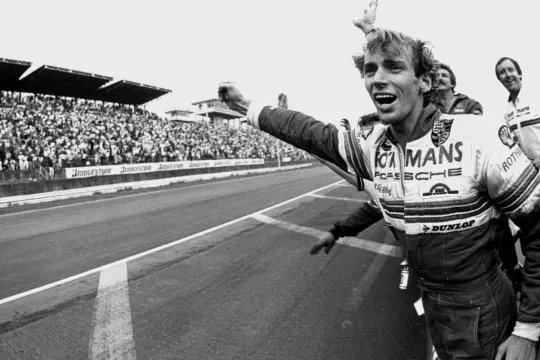 Jubelnder Stefan Bellof im Rennanzug mit "Le Mans" und "Porsche" Aufschrift, jubelnde Menge im Hintergrund.