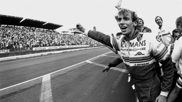 Jubelnder Stefan Bellof im Rennanzug mit "Le Mans" und "Porsche" Aufschrift, jubelnde Menge im Hintergrund.